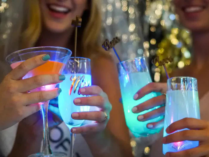 Two people with light skin tone hold fancy looking glasses up to cheers two other glasses. Each glass glows with the Glo Liquid-Activated Cubes.