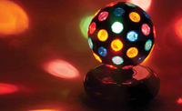 The LAVA Disco Ball sits on the right hand of the picture in a dark room. Small glowing specks of multicolored light reflect off of the walls and floor.