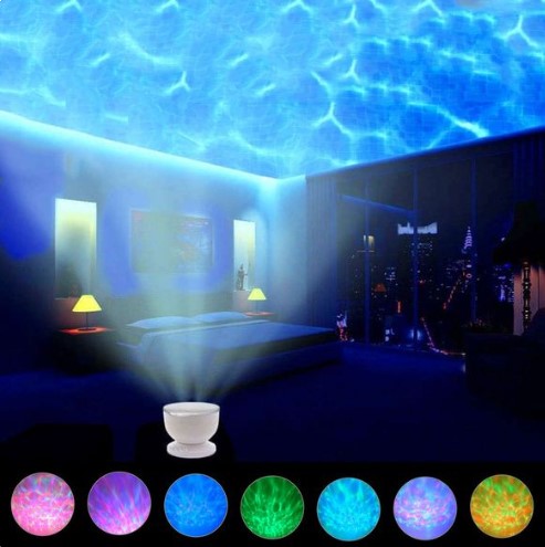 A city apartment bedroom at night, revealing the lit up cityscape out of the window on the right hand side. The Ocean Wave Projector is in the foreground, projecting an illuminated wave pattern on the ceiling in front of the bed.