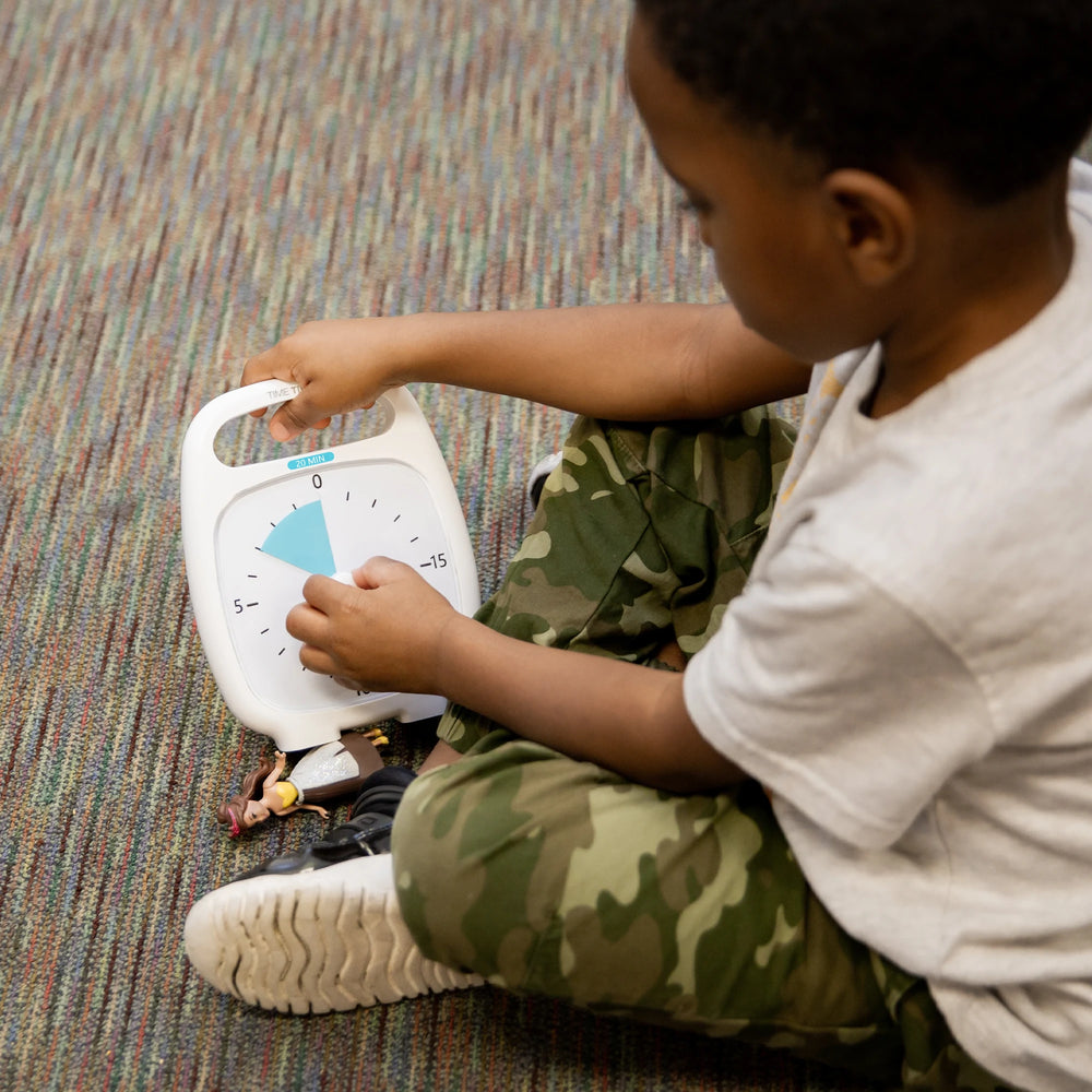 A child with dark skin tone and short black hair is sittingon the ground. They are holding the Time Timer up and turning the center-dial.