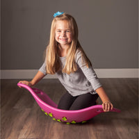 A child with light skin tone and long blonde hair is kneeling on the pink Teeter Popper. They are grabbing either end of the Teeter Popper with their hands and smiling.
