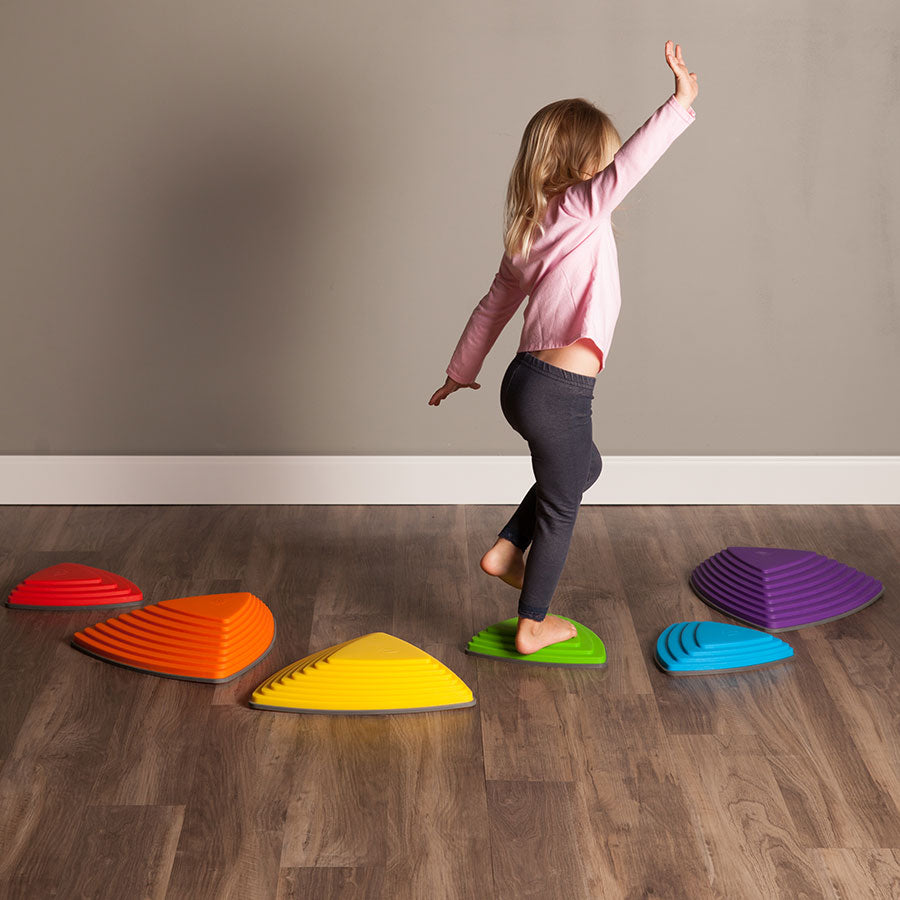 A child with light skin tone and mid-length blonde hair is standing midway through a trail of Gonge Riverstones. Their right leg is balancing on a green Riverstone while their left leg is hovering, and their right hand is above their head while their left hand is down below.