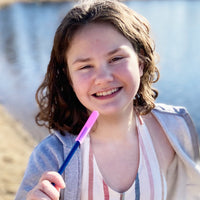 A child with light skin tone and shoulder length wavy brown hair holds the Translucent Pink Krypto-Bite Chewable Pencil Topper in front of their mouth while smiling.
