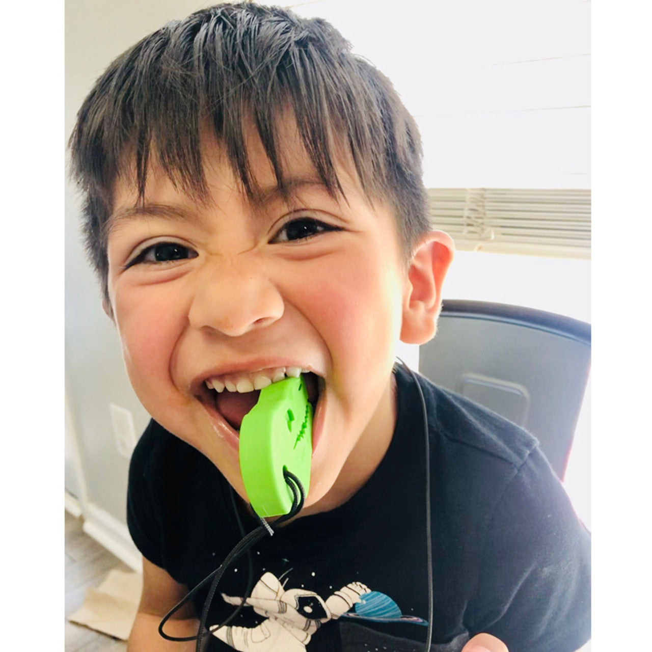 A child with medium light skin tone and short brown hair has the Lime Green XT Dino-Bite Chew Necklace pendant in their mouth. They are smiling while holding it in their teeth.