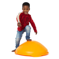 A child with dark skin tone and short black hair is standing over the Gonge Mini Top. Their bare left foot is resting on the top of the Mini Top, the other foot resting on the ground.