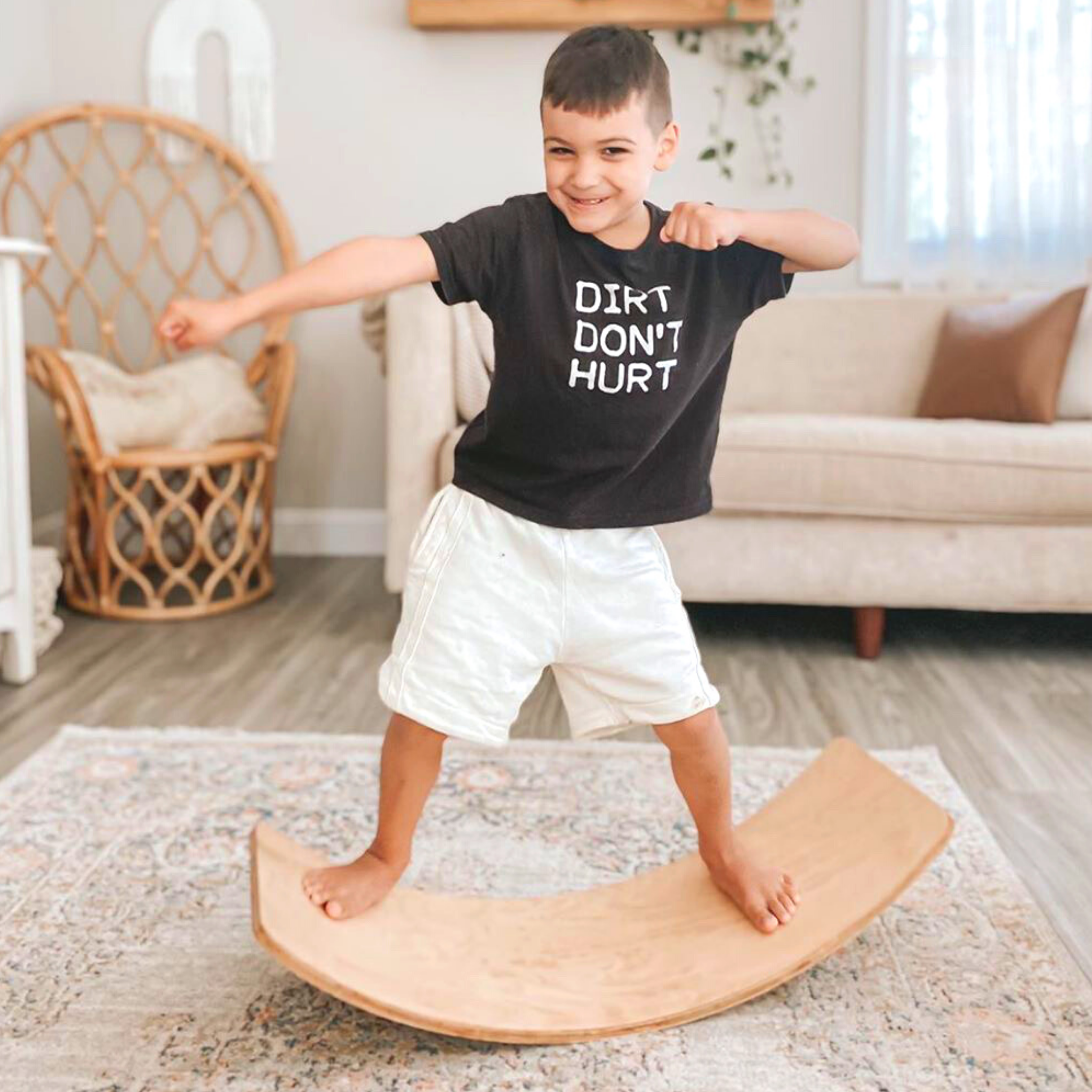 A child with medium light skin tone and short brown hair stands with their feet spread out on either side of the Starter Wobble Board. Their arms are pulled up in a fighting pose and they are smiling.