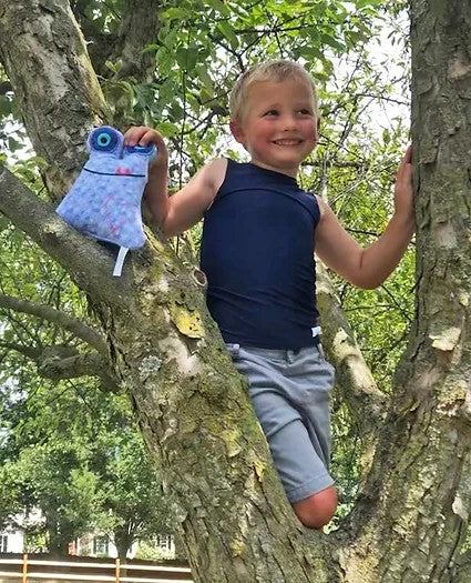A child with light skin tone and short blonde hair is standing in a tree. They are wearing the black Simply Sleeveless Sensory Compression Shirt with grey shorts. They are smiling and holding a blue stuffed animal.