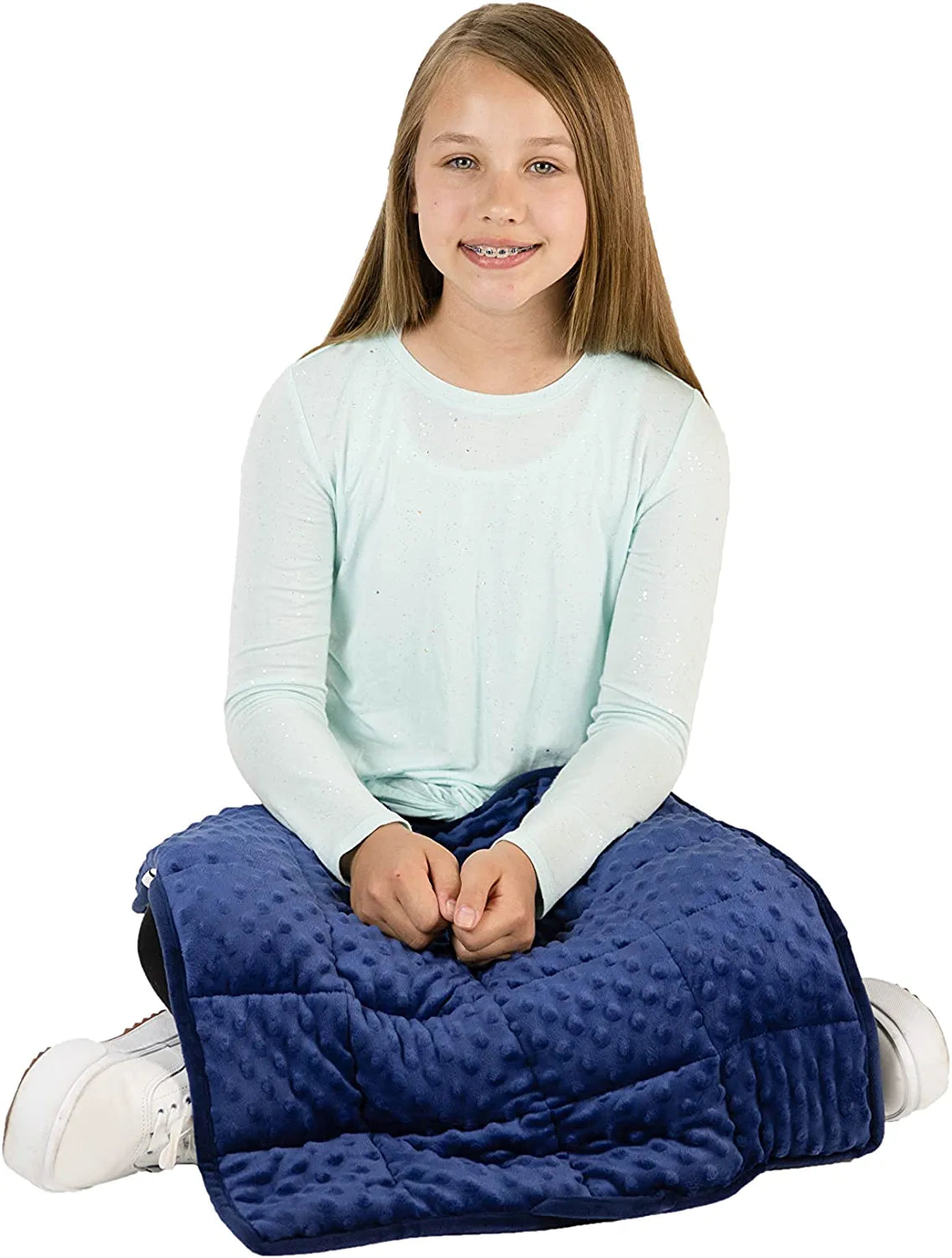 A child with light skin tone and long brown hair sits with their legs criss crossed. A Dual Textured Lap Pad is on their lap, their hands are resting on top of the Pad.