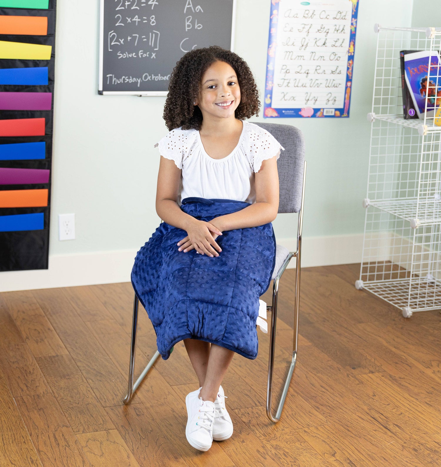 A child with medium skin tone and long tightly curled brown hair sits on a school chair. Their legs and hands are crossed and a Dual Textured Lap Pad is on their lap.