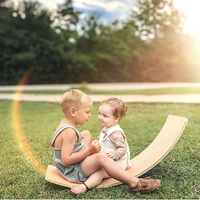 One toddler and one child, both with light skin tone and short hair, are sitting at the edge of a Regular Sized Wobble Board. Their legs are tangled up in each other. The child is blinding while the toddler is looking right at the child.