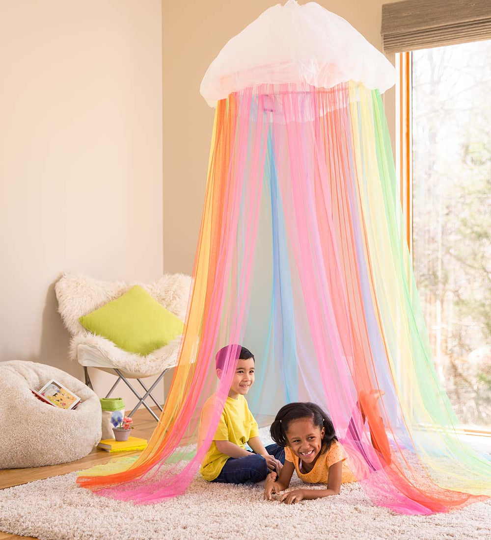 Two children sit underneath the rainbow canopy in a bedroom.