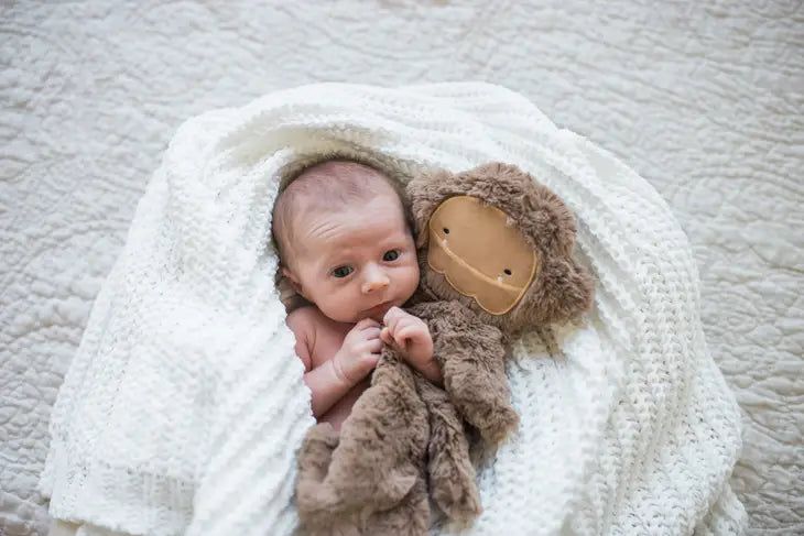 A newborn baby with light skin tone is wrapped in a white blanket. One of its hands is under the Maple Bigfoot Snuggler.