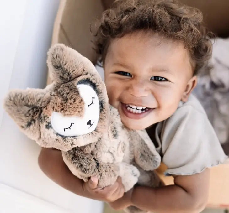 A child with medium skin tone and short curly brown hair smiles big while squeezing Slumberkins Woodland Fox Kin.