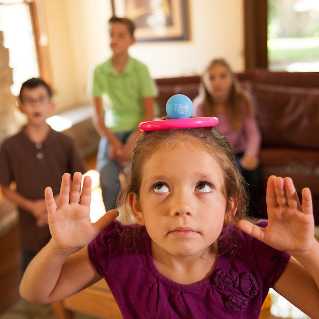 A child with medium light skin tone and brown hair pulled back is hunched over with a blue ball and a pink ring balancing on their head. Their arms are raised and their eyes are looking upward as though concentrating. Three children watch in the background.