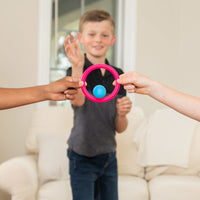 A child with light skin tone and short brown hair is in the background slightly blurry with their hand raised as if throwing an object. Two arms belonging to two different children are holding up a pink ring in the foreground, and there is a blue ball passing through the middle.