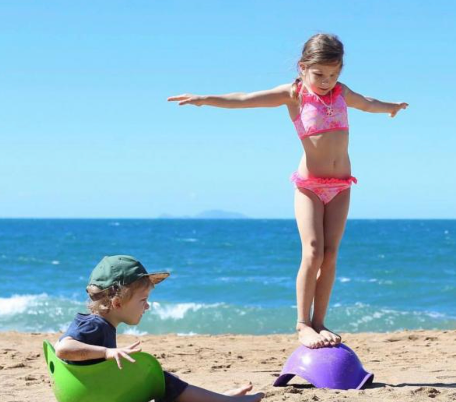 Two children are at a beach on a sunny day. One of the children has short brown hair, is wearing a baseball hat, and is sitting in a green Bilibo. The other child is wearing a pink two piece swimsuit and is balancing on top of an upside down purple Bilibo with their arms spread out as if they are balancing.
