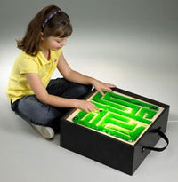 A child with light skin tone and shoulder length brown hair is sitting criss cross applesauce in front of a light table that has the Gel-Maze with Marbles on it. The child is moving the marble through the maze and the table is lit up.