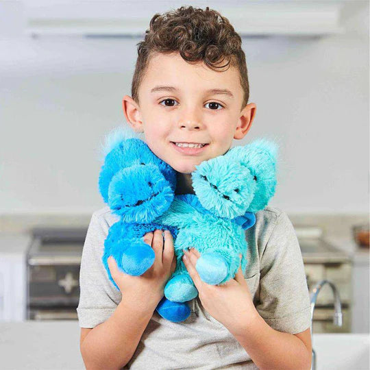 A child with medium light skin tone and short curly hair is smiling and holding both Dinosaur Hugs Warmies up to their chin.