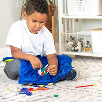 A child with medium skin tone and short brown hair sits criss-cross-applesauce on the floor. The Weighted Lap Pad is on their lap and they are holding a section of intersecting blocks up to assess.