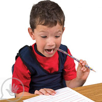 A child with light skin tone and short brown hair sits at a table over a piece of paper. They are putting a Pencil Jaw into their mouth.