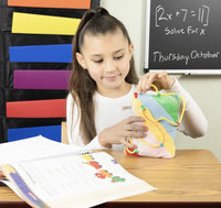 A child with medium light skin and long brown hair in a ponytail on top of their head sits at a desk. They have the Thingamajig Activity Sensory Pillow in their hands, on top of a desk. They are pulling on the cord with one hand and balancing the pillow with the other.