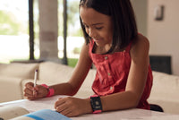A child with medium light skin tone and shoulder length brown hair is sitting at a table and holding a pen. They are wearing the TouchPoints with Wristbands and looking at a piece of paper in front of them.