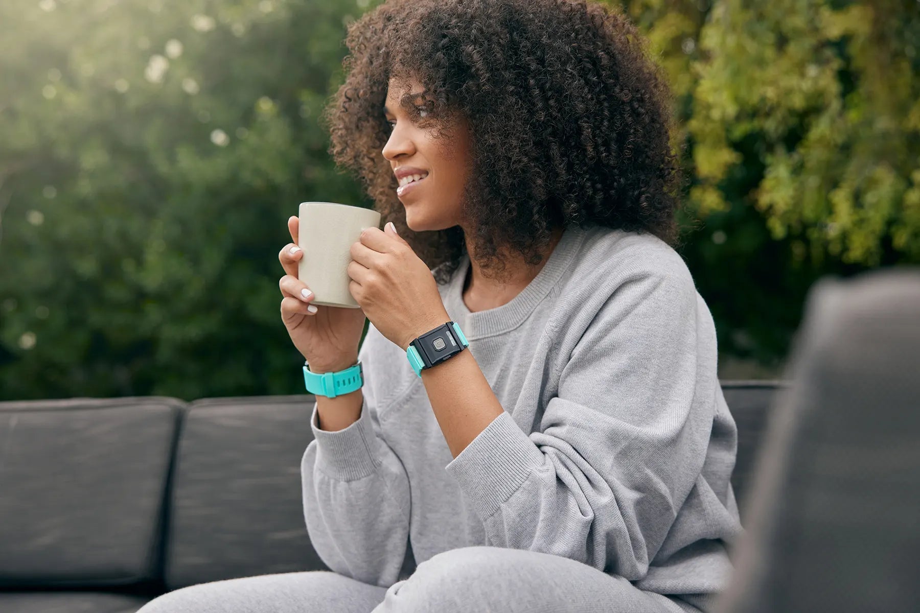 A person with medium skin tone and shoulder length black hair is sitting outside on a couch. They are holding a beverage in a cup and wearing TouchPoints with Wristbands.