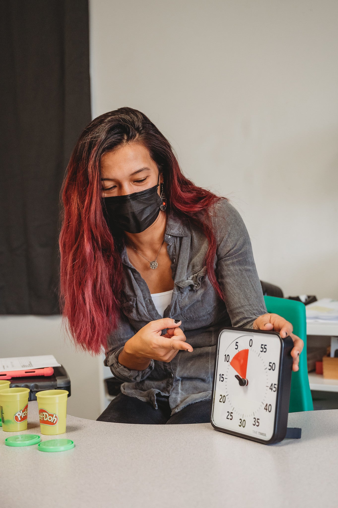 A person with medium skin tone and long dyed red hair is wearing a plain black face mask. They are pointing at the Time Timer Original 8" with one hand and holding it with the other. There are opened cans of Play-Doh on the table.