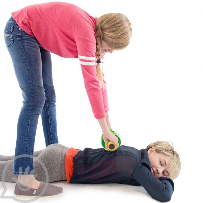 A child with light skin tone and blonde pigtails is bent over another child with light skin tone and short blonde hair lying on their stomach. The standing child is holding a foam roller over the other child's back.