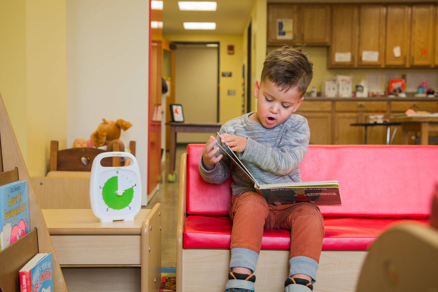 A boy with medium light skine tone and short brown hair is sitting on a child's bench with a book open on their lap. The Time Timer is sitting on a table next to the bench.