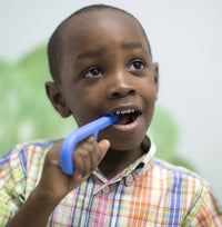 A child with dark skin tone and short black hair is holding the Royal Blue Grabber Original up to their open mouth and has it between their teeth.