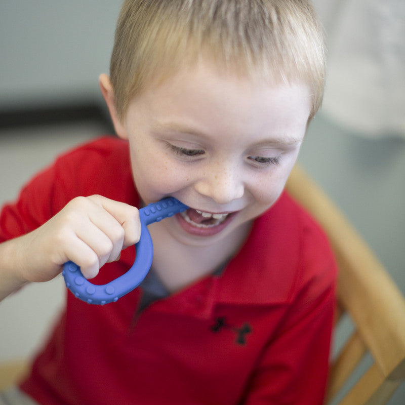 A child with light skin tone and short blonde hair is holding the Royal Blue Textured Grabber up to their open mouth. They have it between their teeth.