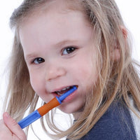 A child with light skin tone and long light brown hair looks into the camera and smiles while holding an orange flavored Senso Brush in their mouth.