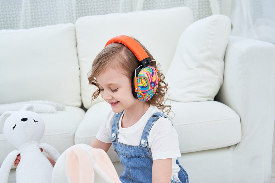 A child with light skin tone and curly, shoulder length brown hair is palying with a white stuffed animal in front of a couch. They are smiling, looking down at the stuffed animal, adn wearing the Lollipop Noise Reduction Headphones.