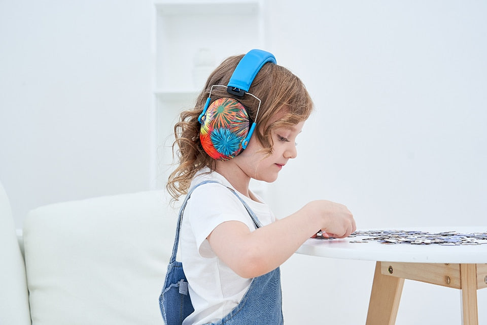 A child with light skin tone and shoulder length, curly, light brown hair is sitting at a table covered in puzzle pieces. They are moving one of the pieces while wearing the Fireworks Noise Reduction Headphones.