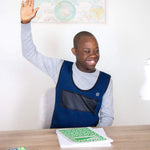 A child with dark skin tone and very short black hair is sitting at a table and raising their hand. They are wearing the Weighted Compression Vest over a long sleeved t-shirt.