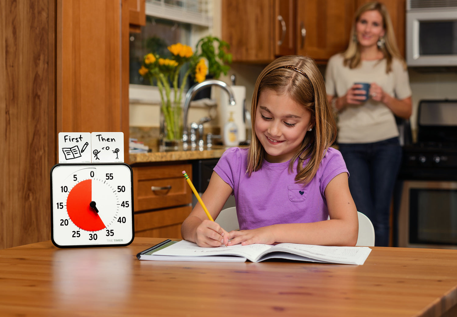 A child with light skin tone and shoulder length blonde hair sits at a desk. They have a booklet open and appear to be doing classwork. The Time Timer Original 8" sits next to them at the table with the Dry Erase Activity Board on top. There is an adult holding a cup of coffee observing them in the background.