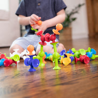 A child's torso sits in the background and holds two Squigz. Several Squigz are connected in the foreground and scattered across a wooden floor.