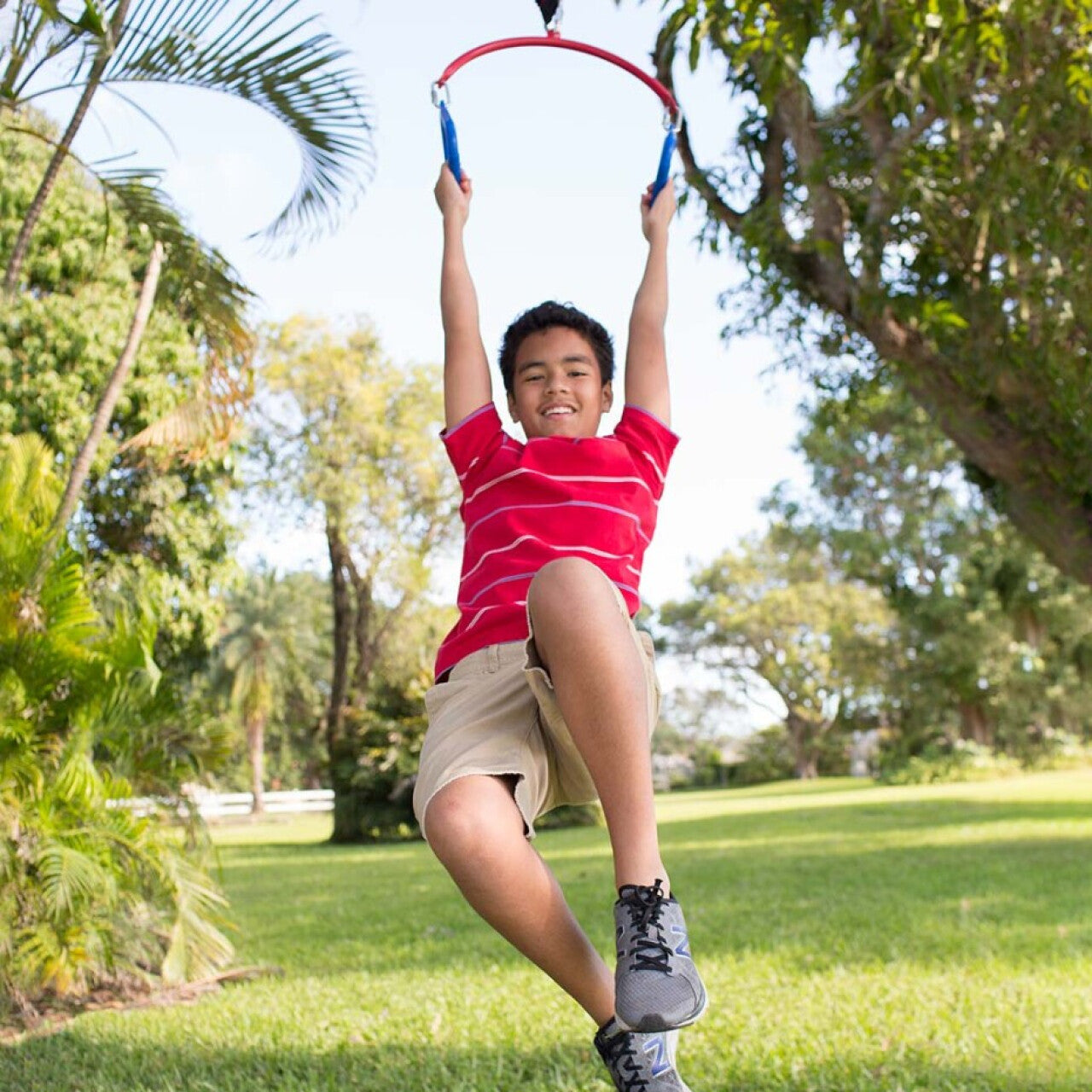A child with medium light skin and short brown hair hangs from the Hanging Rings. They swing forward as they dangle.