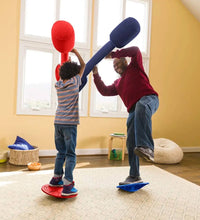 A child and an adult are standing on two separate balance boards. The adult only has one foot on their balance boarrd and is holding the Joust in a defensive position.