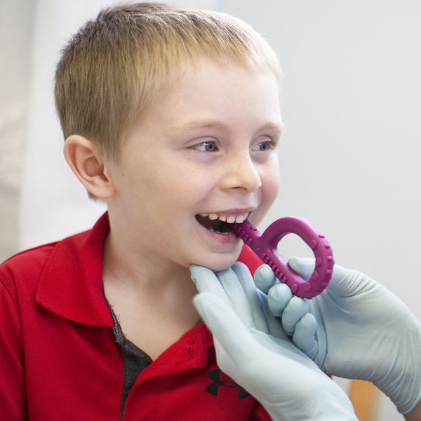 A child with light skin tone and short blonde hair is smiling at someone off camera. Two gloved hands are holding the magenta Textured Grabber up to their mouth, and they are closing their teeth around it.