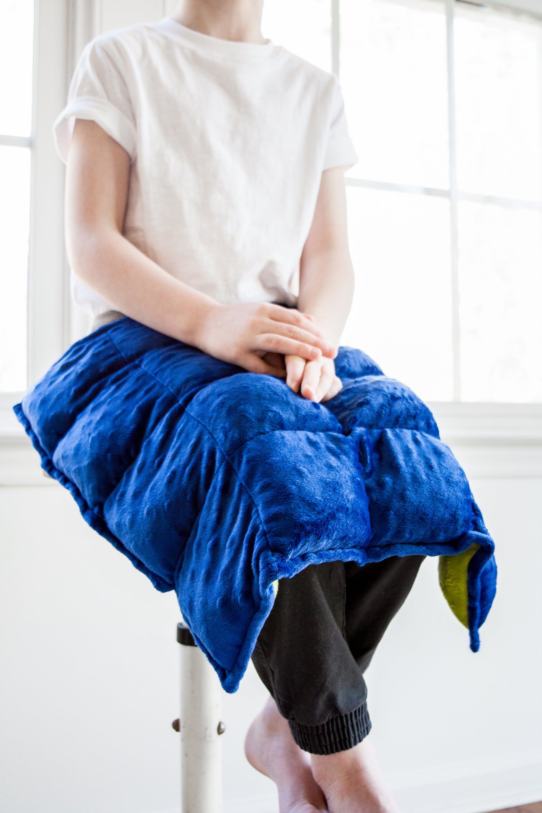 A child with light skin tone sits on a stool with a Weighted Lap Pad on their lap.