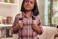 A child with medium skin tone and shoulder length brown hair is wearing a pink backback and shirt. There are TouchPoints attached to both backpack straps.