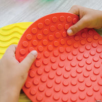Two hands with medium skin tone hold up the red dotted Sensory Mat.