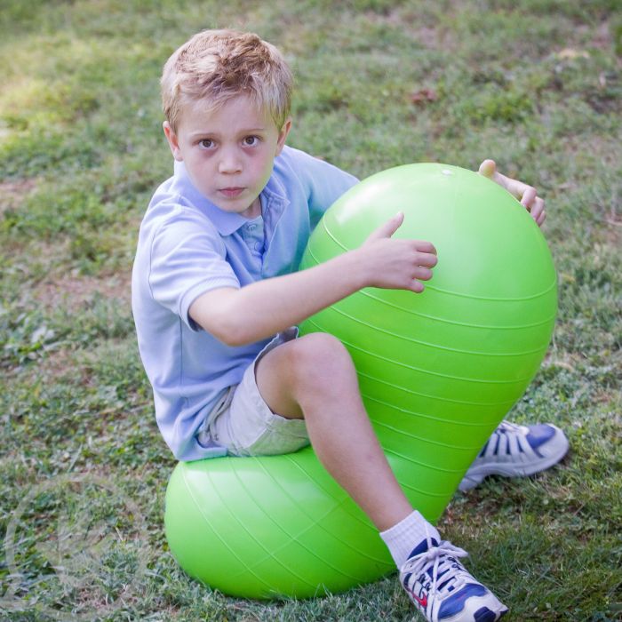 A child with light skin tone and short blonde hair sits on a green Peanut Ball in dry grass.