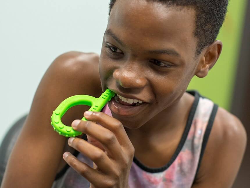 A child wtih dark skin tone and short black hair holds the Lime Green Textured Grabber up to their mouth.  They have it in between their teeth.