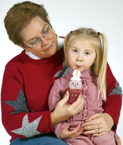 A child with light skin tone and two blonde pig tails is sitting on the lap of an adult with light skin tone and short brown hair. The adult is holding Sippin' Bear up to the mouth of the child while they sip from the straw.