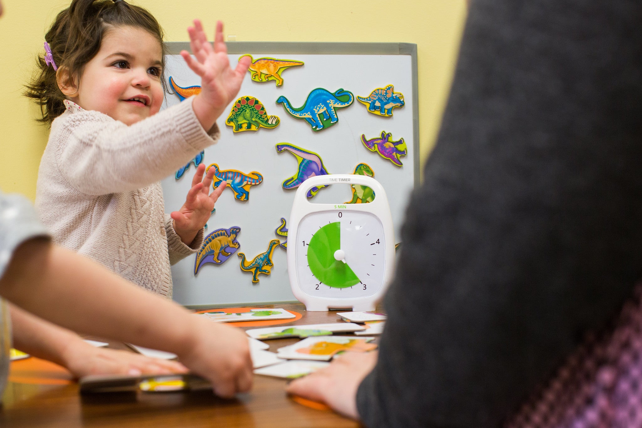A child with light skin tone and short brown hair is standing in front of a dry erase board covered in magnetic dinosaurs. The child appears to be participating in a group activity. The Time Timer is set at 3 minutes and standing in front of the dry erase board.