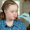 A child with light skin tone and french braided brown hair looks down as a gloved hand approaches their mouth with a Preefer Tip attached to a Z-Vibe.