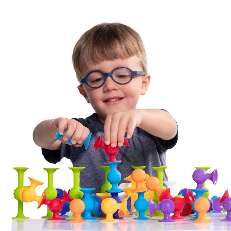 A child with light skin tone and short light brown hair wears blue glasses.  They are holding a red Squig on a short stack of Squigz, and trying to connect it to a blue Squig. 